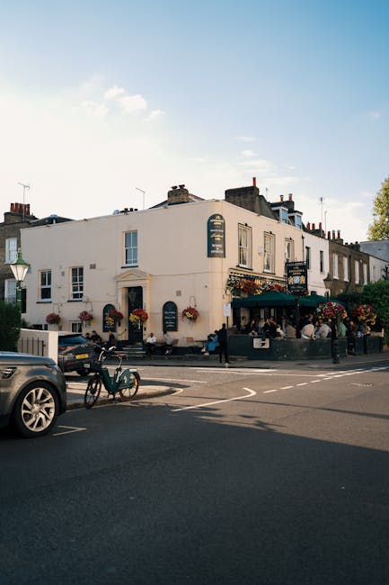 Photograph of a two-story building situated on a street corner during daytime, featuring white exterior walls and multiple windows. The ground floor appears to be a pub or restaurant, indicated by out