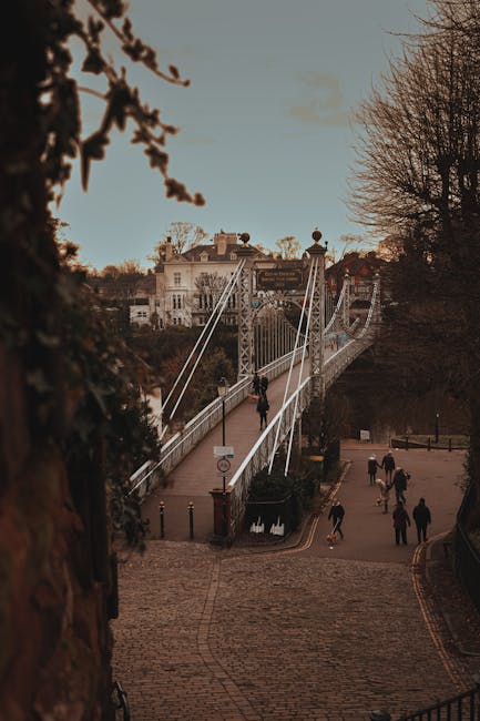 A view of Hammersmith Bridge spanning across the River Thames under a cloudy sky, with the bridge's white suspension structure and tall towers visible. On the riverbanks, there are modern residential 